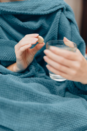 Woman in blue robe holding glass of water and pill while wrapped in blanket at homeの写真素材