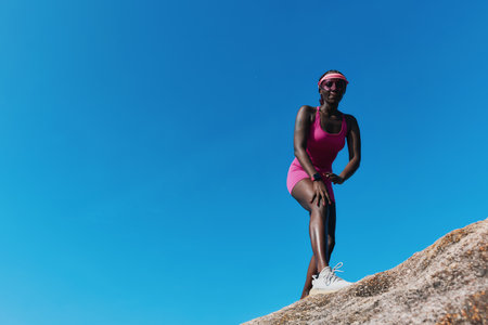 A fit woman in a pink workout outfit stands on a rocky surface under a clear blue sky, demonstrating an active lifestyleの写真素材
