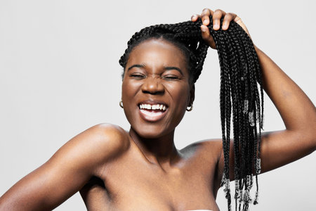 Happy young woman with braids playfully pulling her hair, joy and confidence expressed against a...