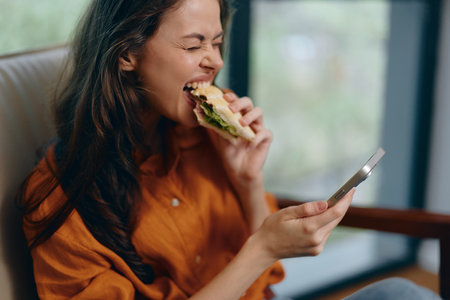Young woman enjoying a delicious sandwich while texting on her phone, showcasing a relaxed vibe and healthy eating habits in a cozy indoor settingの写真素材