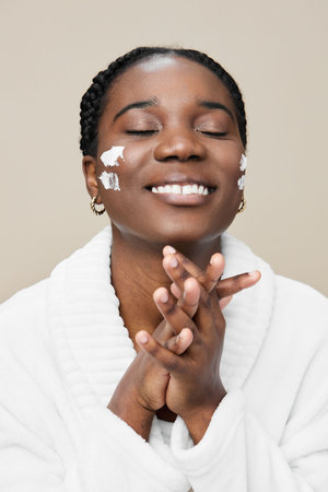 Joyful African woman applying skincare cream, radiating happiness and self care, in a white bathrobe against a soft beige backgroundの写真素材