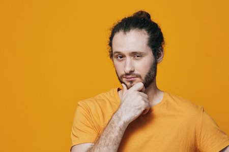 Thoughtful young man with a beard in an orange t shirt against a bright yellow background, contemplating and reflecting with a confident expressionの写真素材