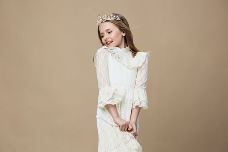 Charming young girl wearing a white dress and a crown, smiling joyfully against a beige background, exuding innocence and eleganceの写真素材