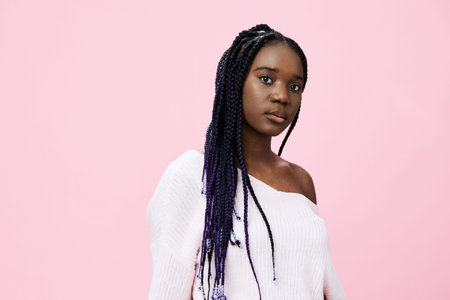 Portrait of a young Black woman with braids, wearing a cozy white sweater against a pink background, expressing confidence and eleganceの写真素材