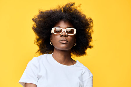 Stylish young woman with curly hair wearing sunglasses, posed against a vibrant yellow background,...