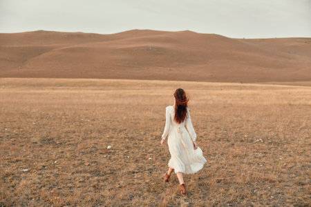 Woman in white dress walking through open field with hills in background, embracing the beauty of nature and freedomの写真素材