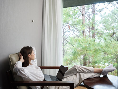 Relaxed young woman enjoying a peaceful moment at home, working on her laptop, surrounded by nature through large windows, promoting a serene lifestyleの写真素材