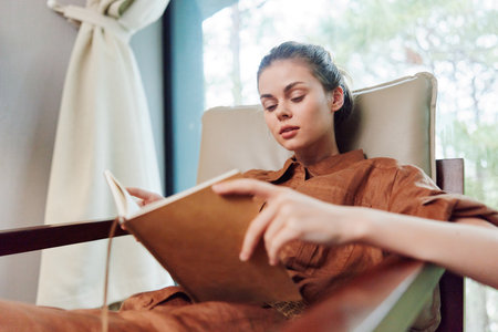 Woman reading a book in a cozy chair, dressed in a brown outfit, displaying calm and focused emotions, with a natural green background, reflecting relaxation and tranquilityの写真素材