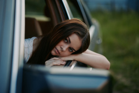 A reflective young woman resting her head on the car window, showcasing emotions of contemplation,...