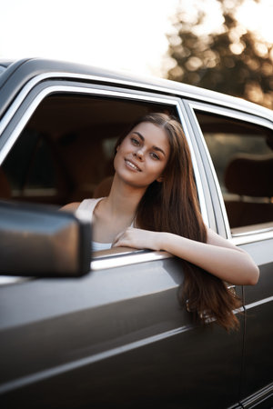 Young woman smiling in a car window during sunset, showcasing carefree emotions and a casual style,...