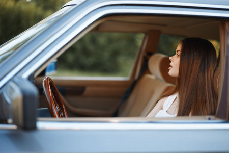 Thoughtful young woman with long hair sitting in a vintage car, gazing out the window, reflecting on...