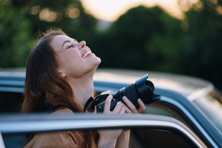 Young smiling woman holding a camera, capturing a moment outdoors, with a joyful expression in a natural setting during golden hour.の写真素材