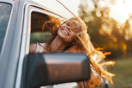Young woman with flowing hair smiles joyfully from a car window during sunset, showcasing freedom...