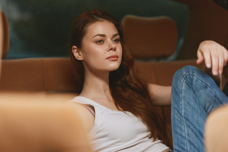 Elegant young woman with long hair in casual outfit relaxing in the backseat of a car, conveying a...