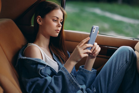 Young woman in casual denim outfit using smartphone in car, showcasing modern lifestyle and urban...