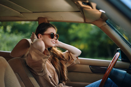 Young woman enjoying the sun in a car, wearing sunglasses and smiling, with a lush green background, capturing a feeling of freedom and joy during summer.の写真素材
