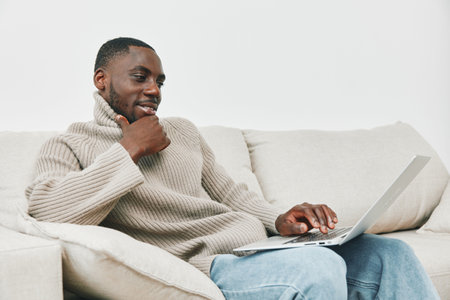 Young African man in cozy sweater using laptop at home, showcasing comfort and modern lifestyle, focusing on work and relaxation in a contemporary settingの写真素材
