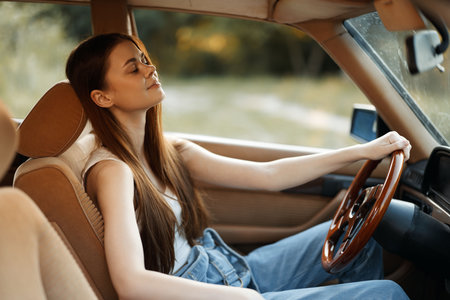 Young woman enjoying a peaceful moment in a vintage car, with sunlit nature in the background, wearing casual blue jeans and a relaxed expression, evoking freedom and serenity.の写真素材