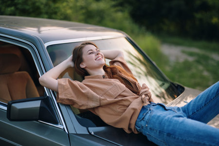Young woman relaxing on a car roof in casual clothing, smiling joyfully amidst a lush green background, capturing a carefree summer vibe.の写真素材