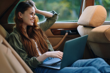 Young woman working on a laptop in a car, smiling and relaxed, showcasing a casual outfit and warm...