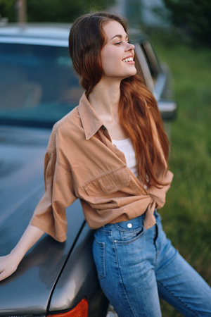 Young woman smiling in casual outfit, enjoying summer outdoors by a vintage car with a blurred green background and warm light.の写真素材