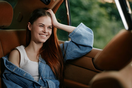 Happy young woman with long hair enjoying a road trip in a vintage car, wearing a denim jacket and...