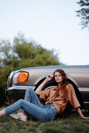 Stylish young woman with long hair sitting next to a vintage car in a natural setting, wearing a brown top and jeans, evoking a carefree summer vibe.の写真素材