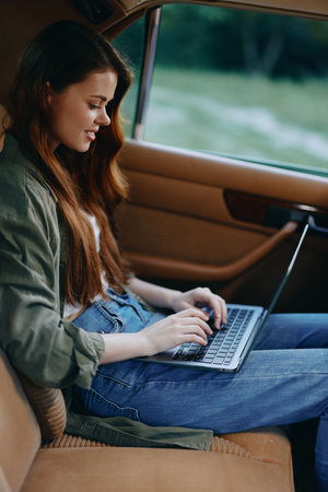 Young woman working on a laptop in a car, wearing casual clothing, with a relaxed expression,...