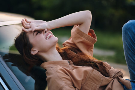 Young woman laughing joyfully while resting on a car hood, dressed in casual clothing, with a vibrant green background, evoking a carefree summer mood.の写真素材