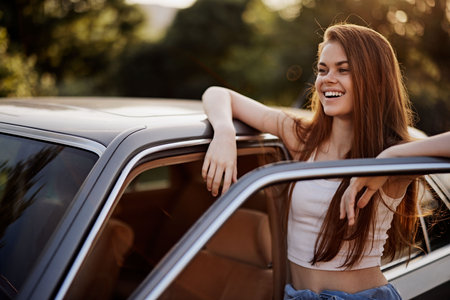 Smiling young woman leaning on a car, enjoying a sunny day, wearing casual summer clothes, and surrounded by a lush green background.の写真素材