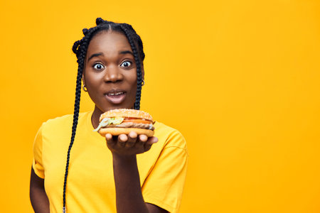 Happy Black woman holding a delicious burger against a vibrant yellow background, showcasing...