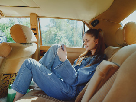 Young woman in casual denim outfit enjoying her smartphone in a cozy car interior, expressing happiness and relaxation during a sunny day trip.の写真素材