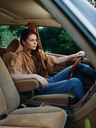 Stylish young woman in a vintage car, wearing casual attire, expressing a thoughtful mood amidst a natural green backdrop.の写真素材