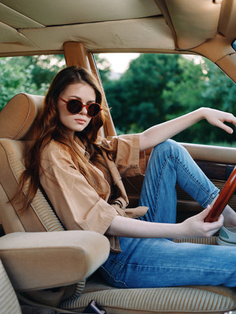 Young woman in sunglasses sitting in a vintage car, wearing a casual outfit, showcasing a relaxed summer vibe with greenery in the background.の写真素材