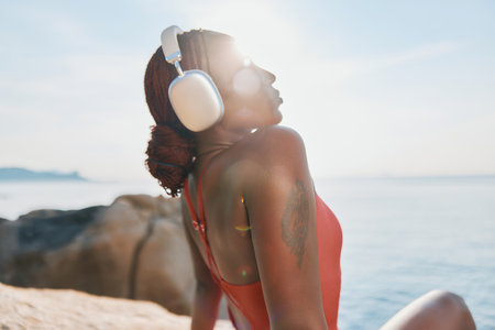 Woman enjoying music by the sea at sunset, wearing headphones, with a serene expression and sun reflecting behind herの写真素材