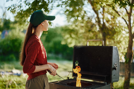 Young woman in a green cap grilling outdoors in a backyard during summer, enjoying the vibrant atmosphere with flames and greeneryの写真素材