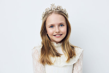 Charming young girl with a crown, wearing a vintage white dress, smiling sweetly, against a light gray background, perfect for themes of childhood and innocenceの写真素材