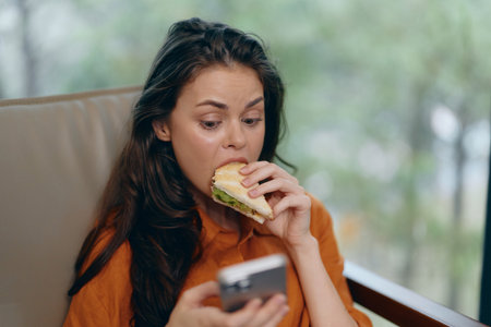 Young woman enjoying a sandwich while scrolling on her phone in a cozy cafe environment, reflecting...