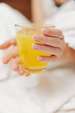 Woman holding a glass of orange juice while laying on a bed with pillows and a blanket on top of herの写真素材