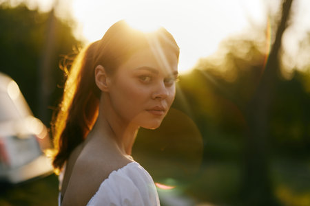 Young woman with long hair in casual white dress, softly illuminated by sunset, captures a moment of contemplation and serenity in natureの写真素材
