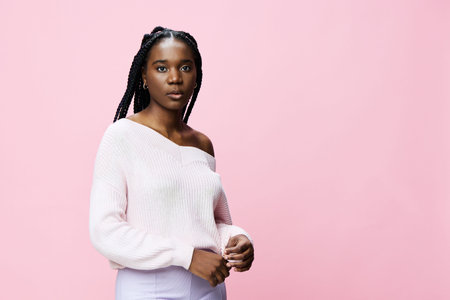 Elegant young black woman in a soft pink sweater poses against a pastel pink background, conveying confidence and style through her calm expressionの写真素材