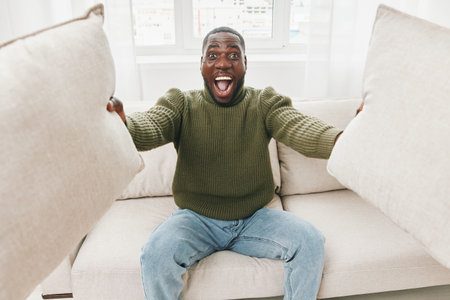 Happy young Black man expressing joy while sitting on a sofa, holding decorative pillows in a cozy living room setting with soft lightingの写真素材
