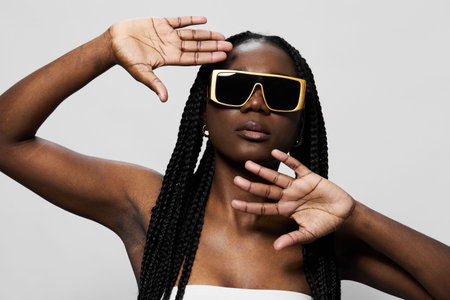 Confident young woman with braids wearing oversized sunglasses against a minimalistic background, showcasing modern fashion and bold styleの写真素材