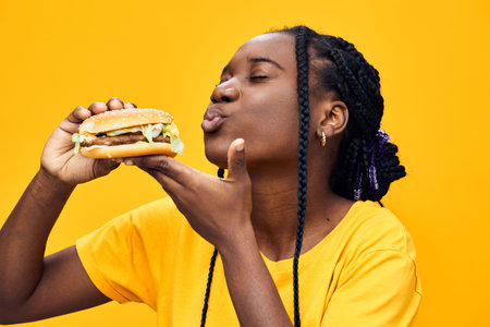 Happy young woman with braids enjoying a delicious burger against a vibrant yellow background,...