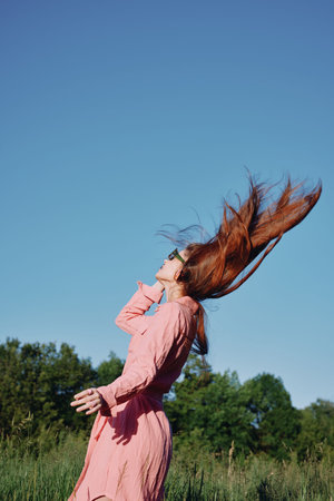 A joyful young woman with long red hair, dressed in a pink dress, enjoying a sunny day outdoors against a clear blue sky in a lush green fieldの写真素材