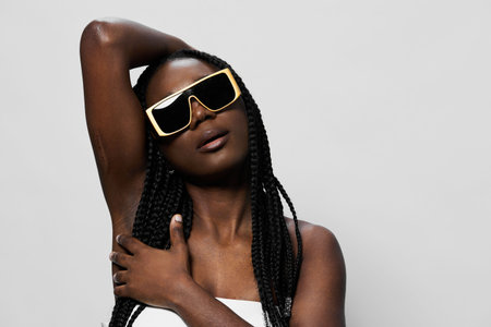 Elegant African woman wearing trendy sunglasses poses confidently, showcasing her braided hair and stylish outfit against a neutral backgroundの写真素材
