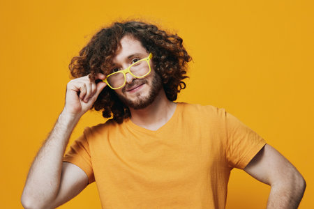 Cheerful young man with curly hair, wearing yellow glasses and a bright orange t shirt against a vibrant yellow backgroundの写真素材