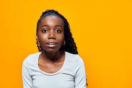 Young Black woman with curly hair smiling against a bright yellow background, showcasing joy and confidence in casual grey attireの写真素材