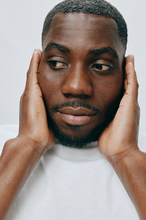 Portrait of a thoughtful man with dark skin showing emotion while touching his face in a minimalist setting, wearing a plain white shirt against a light backgroundの写真素材