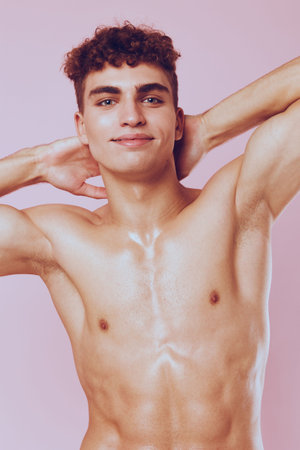 Confident young man with curly hair posing shirtless, showcasing his toned physique against a pink background. Fitness and self-expression concept.の写真素材